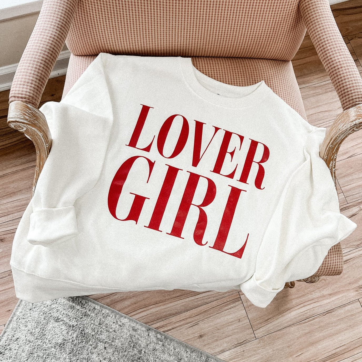 A white crewneck sweatshirt with the text 'Lover Girl' printed in red text on the front, placed on a chair in a cozy living room setting.