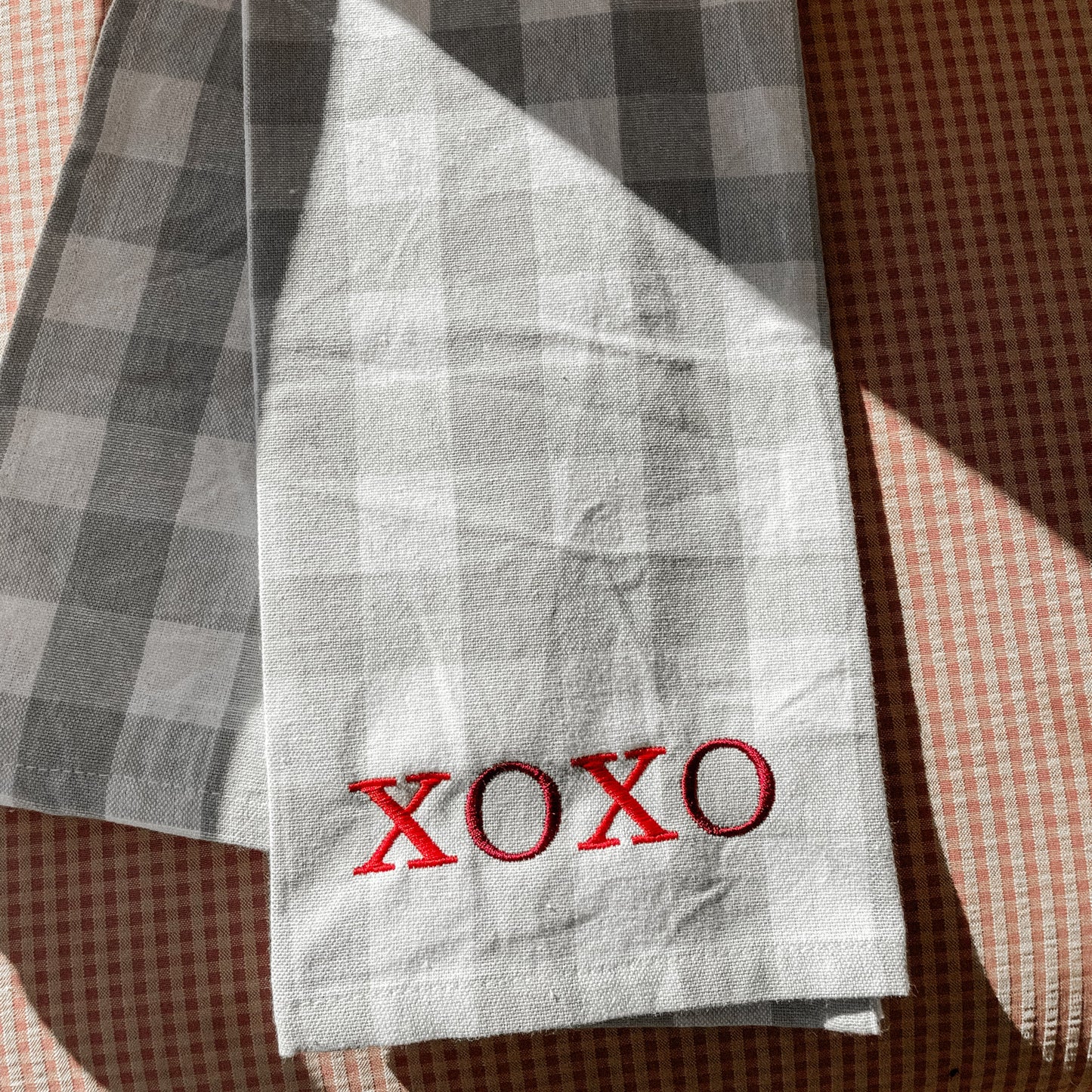 A folded white checkered kitchen towel with the word 'XOXO' printed in red on the front, placed on a checkered patterned surface.