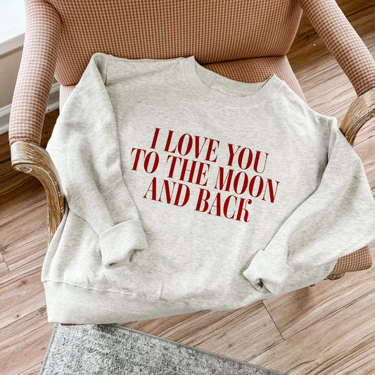 A gray crewneck sweatshirt with the text 'I love you to the MOON and BACK' printed on the front, placed on a chair in a cozy living room setting.