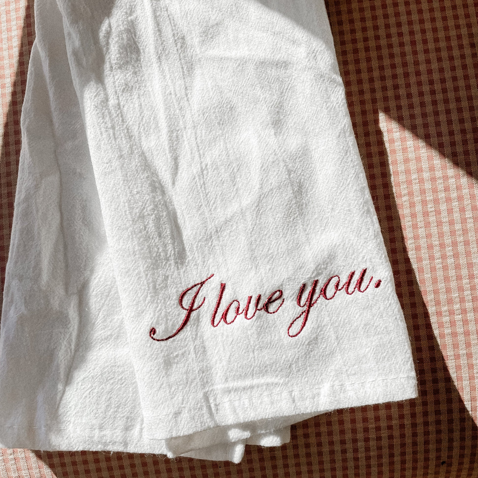 A white kitchen towel with the phrase 'I love you' embroidered in red thread on it, placed on a surface with a checkered pattern.