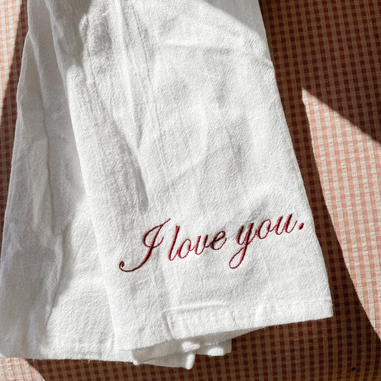 A white kitchen towel with the phrase 'I love you' embroidered in red thread on it, placed on a surface with a checkered pattern.