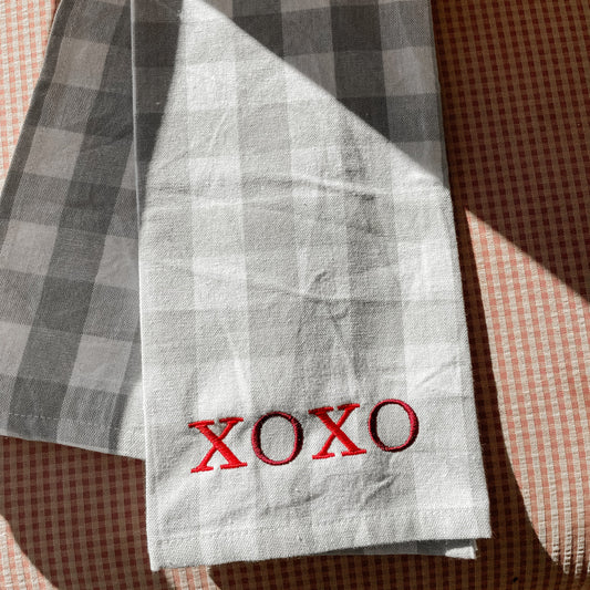 A folded white checkered kitchen towel with the word 'XOXO' printed in red on the front, placed on a checkered patterned surface.